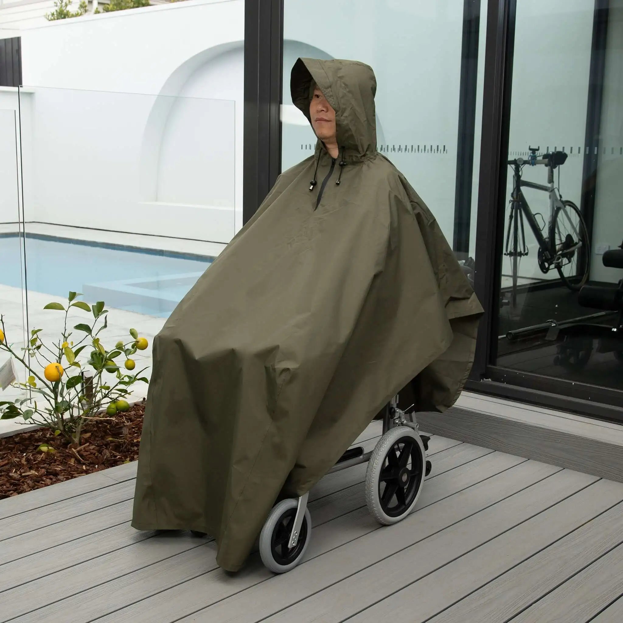 Person seated in a wheelchair wearing a waterproof wheelchair poncho in olive green. Designed for full-body rain protection, this poncho features a hood with adjustable drawstrings and a roomy fit to cover both the user and wheelchair. Made from lightweight, durable, and water-resistant fabric, it ensures comfort and mobility while shielding against wind and rain. Ideal for outdoor use, travel, and everyday weather protection.