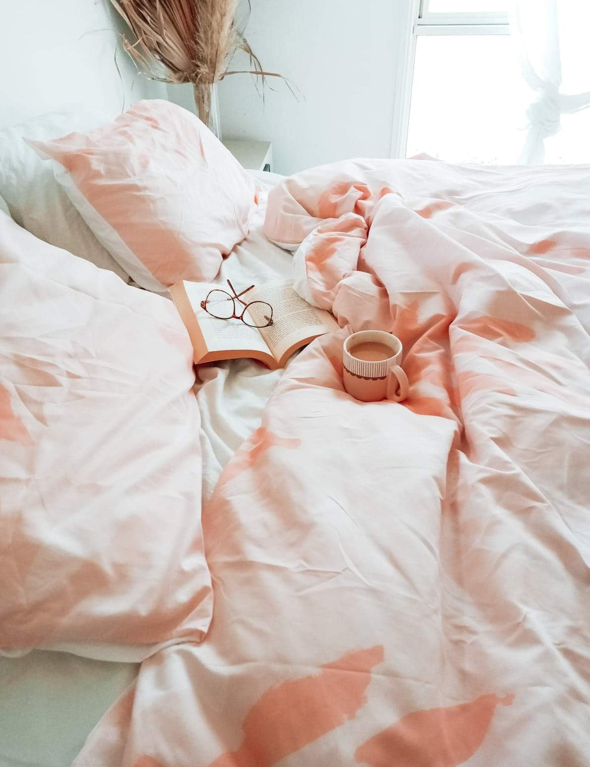 The Botanic quilt cover in Peach on a bed with a cup of coffee, a book & reading glasses sitting onto of the cover.
