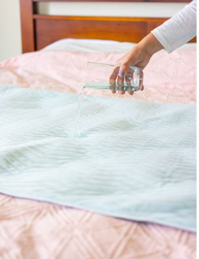 Boss 40 bed pad, on top of a bed with a lady's hand pouring liquid on it to demonstrate its waterproof and absorbent qualities
