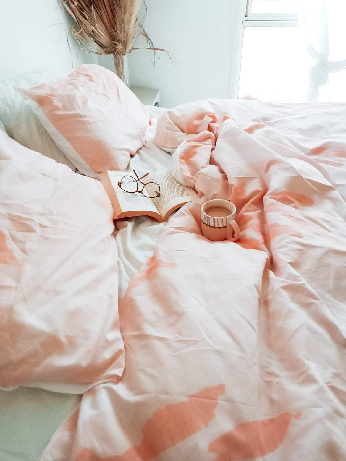 The Botanic quilt cover in Peach on a bed with a cup of coffee, a book & reading glasses sitting onto of the cover.