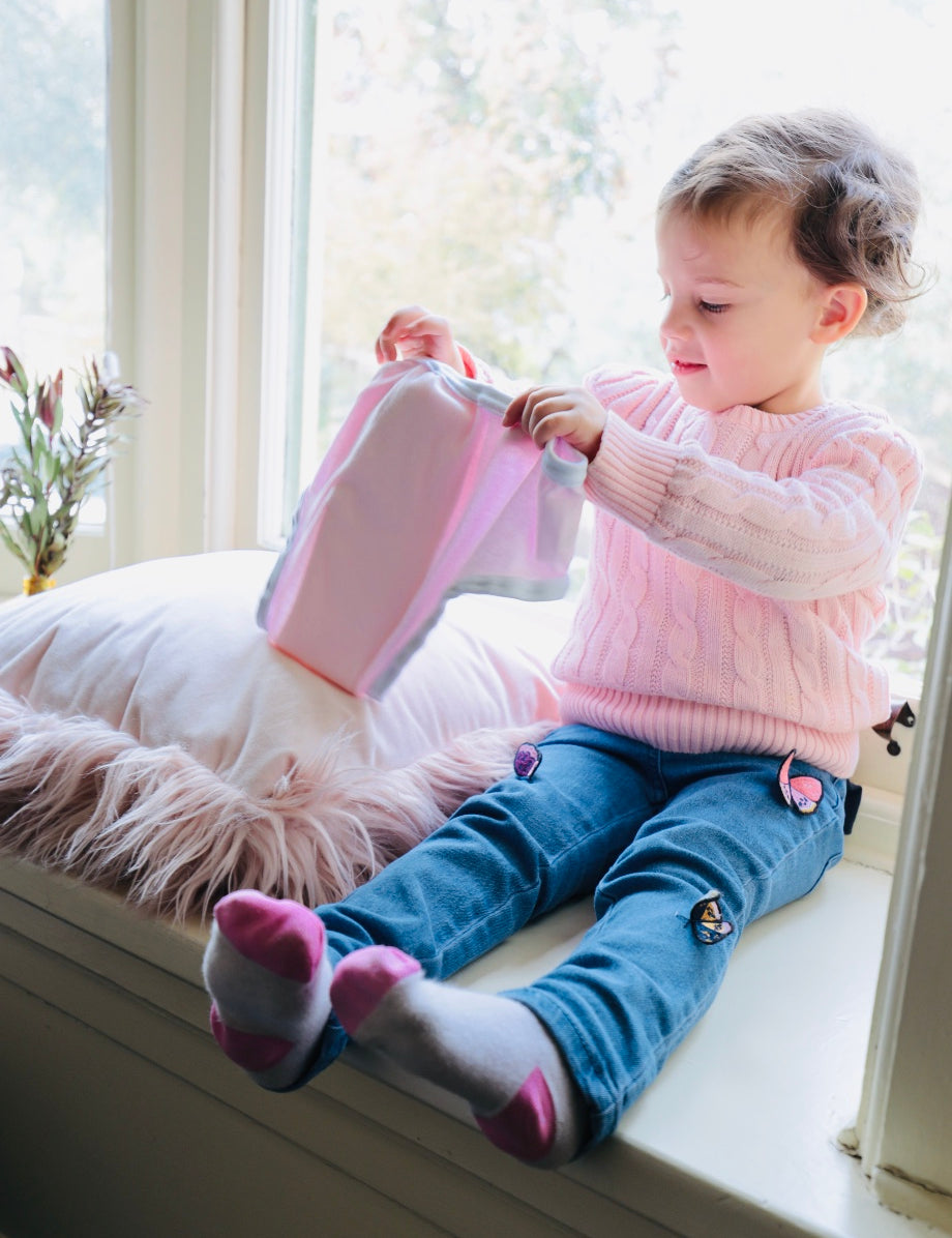 Toddler girl in a pink jumper sitting by a light filled window holding a pair of pink incontinence briefs