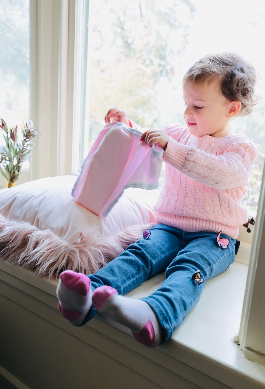 Toddler girl in a pink jumper sitting by a light filled window holding a pair of pink incontinence briefs